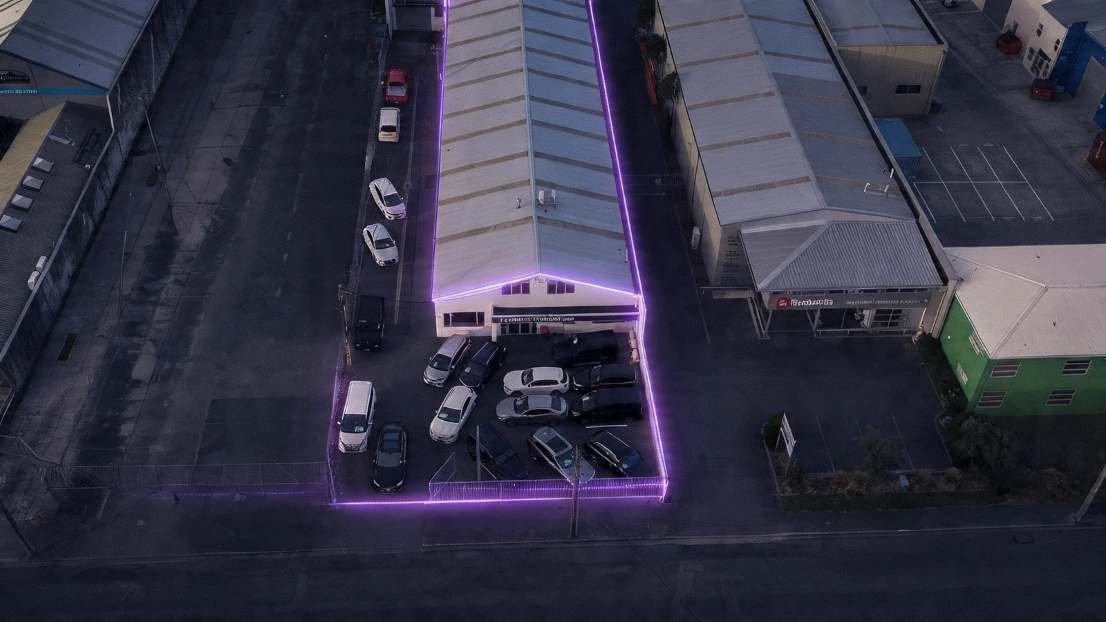 Premium Wholesale Cars Christchurch dealership exterior at dusk with a cloudless indigo to rose gradient sky, warm amber interior light glowing from showroom windows and upstairs offices, and the brand purple car icon and signage illuminated